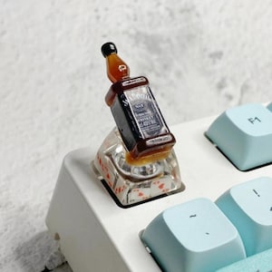 Könnte beinhalten: Ein Miniatur-Whiskeyflaschen-Keycap in Braun und Schwarz. Der Keycap ist auf einer transparenten Basis mit roten Herzsymbolen montiert und liegt auf einer weißen Tastatur.