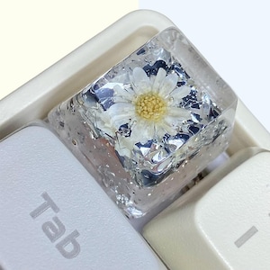 Könnte beinhalten: Eine benutzerdefinierte Tastenkappe für eine Tastatur mit einer weißen Gänseblümchenblüte, die in klarem Harz mit silbernem Glitzer gegossen ist.