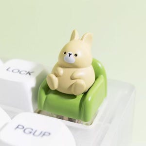 May include: A light yellow bunny figurine sitting on a small green chair, designed as a keyboard keycap. The bunny has black eyes and a white nose. The keyboard keys are white, with the words "LOCK" and "PGUP" visible.