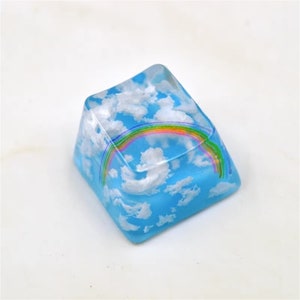 May include: A clear blue and white resin keycap with a rainbow design. The keycap features a cloudy sky with a rainbow arching over it.
