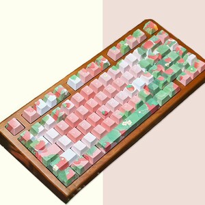 May include: A wooden keyboard with pink and white keycaps featuring a strawberry and leaf design. The keyboard has a wooden frame and a green and white pattern on the keys.