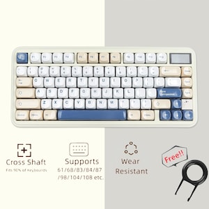 Pode incluir: Um teclado mecânico creme com teclas azuis e bege. O teclado tem um layout compacto com teclas de função e um visor digital. A imagem também mostra um extrator de teclas e texto que diz "Cross Shaft", "Supports" e "Wear Resistant".