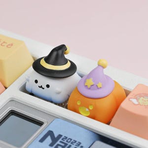 May include: Two custom mechanical keyboard keycaps shaped like characters. One is a white ghost wearing a black witch hat with a yellow band. The other is an orange pumpkin wearing a light purple wizard hat decorated with two yellow stars.