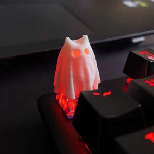 May include: A white, ghost-shaped mechanical keyboard keycap featuring two circular cutouts for eyes. The keycap is mounted on a switch that emits a bright red light, which illuminates the interior of the ghost figure and shines through the eye holes.