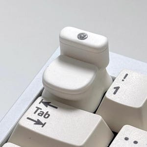 Könnte beinhalten: Eine neuartige, weiße Toiletten-Tastenkappe mit einem kleinen, dekorativen Edelstein oben, platziert auf einer weißen Tastatur. Die Tastenkappe ersetzt die Standardtaste. Die "Tab"-Taste ist sichtbar.