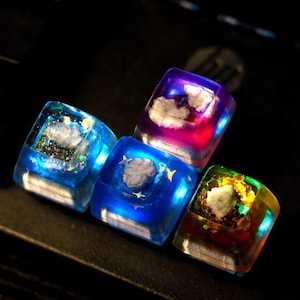 Könnte beinhalten: Vier durchscheinende, quadratische Tastenkappen mit Wolkenmotiven. Die Tastenkappen sind in Blau-, Lila- und Goldtönen gehalten und mit Glitzer- und Sternakzenten versehen. Die Tastenkappen sind auf einer dunklen Oberfläche angeordnet.