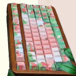 May include: A wooden keyboard with a strawberry and leaf design. The keys are pink and white with a light brown wooden frame.