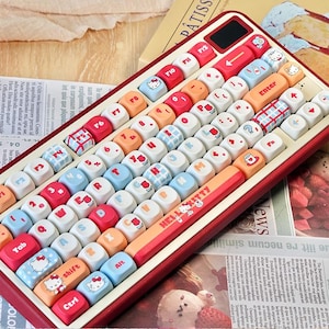 May include: A mechanical keyboard with a deep red frame and a variety of colourful keycaps. The keys feature pastel blue, orange, white, and red colours with printed icons of Hello Kitty, flowers, and plaid patterns. The spacebar displays the text HELLO KITTY.