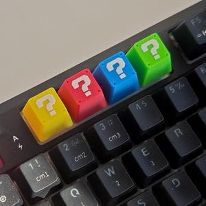 May include: A close-up of a black keyboard with four colorful question mark keycaps. The keycaps are yellow, red, blue, and green. The question marks are white. The keyboard has other black keys with white lettering.