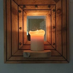 May include: A wooden frame with a mirror inside. The frame is decorated with small metal studs. A lit candle sits on a wooden shelf in front of the mirror.