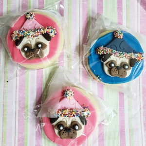 May include: Three decorated sugar cookies with a pug face and a party hat on top. The cookies are pink, blue, and pink with colorful sprinkles around the edges.