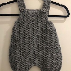 May include: A gray, handmade crochet romper for a baby, featuring a textured stitch pattern. The romper has button closures on the straps and a rounded bottom. It is displayed on a black hanger against a white wall.