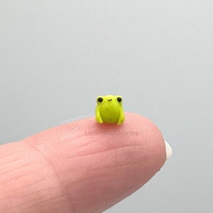 May include: A tiny, bright yellow frog figurine with black eyes sits on a finger. The frog is very small, about the size of a fingernail.
