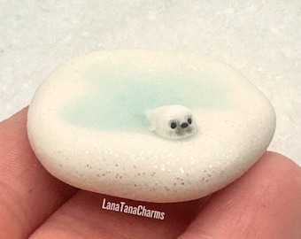 May include: A miniature, handcrafted charm featuring a white seal swimming in a light blue pool. The seal has black eyes and is set in a white, glittery, oval-shaped base. The charm is held in a hand.