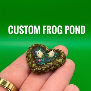 May include: A heart-shaped miniature frog pond with two white frog figurines floating in a glittery blue and purple pond. The pond is surrounded by brown and green textured material. The text "CUSTOM FROG POND" is above the pond.