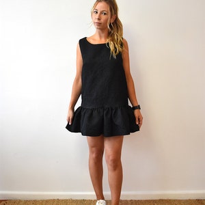 May include: A black sleeveless dress with a ruffled hem. The dress is made of a textured fabric and has a square neckline. The model is wearing white sneakers.