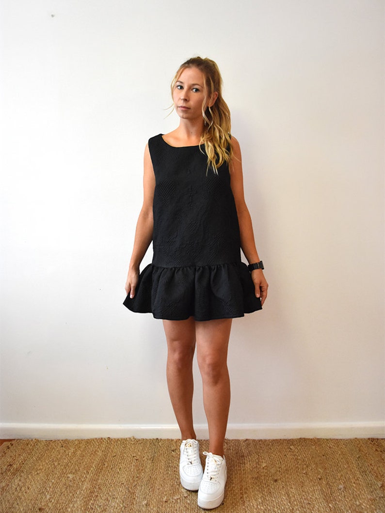 May include: A black sleeveless dress with a ruffled hem. The dress is made of a textured fabric and has a square neckline. The model is wearing white sneakers.