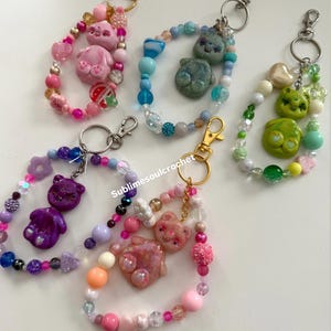 May include: A collection of beaded keychains featuring bear-shaped charms in various colors. Each keychain has a lobster clasp and is adorned with an assortment of beads in shades of pink, purple, blue, and green. The text "Sublimesoulcrochet" is visible.