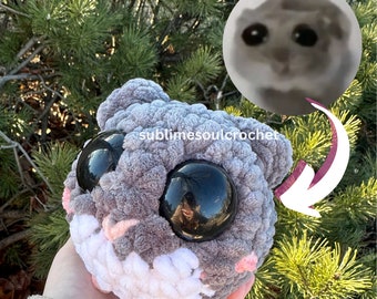 May include: A gray and white crocheted hamster plush toy with large black eyes. The toy is being held in a hand and has a pink nose and mouth. The text "sublimesoulcrochet" is visible on the toy.