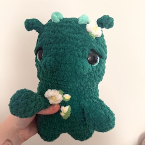 May include: A handmade, plush creature in a deep green hue, adorned with small white and yellow flower accents. The creature has large, expressive eyes and small, rounded ears. The soft, textured material suggests a cuddly toy or decorative item.