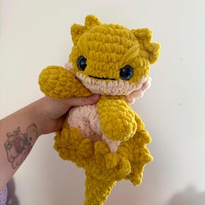 May include: A handmade crochet lizard plushie in yellow and pale pink. The toy has large, sparkly blue eyes and a smiling expression. The lizard has a spiky dorsal ridge and a long tail, suitable for a child's toy or nursery decoration.