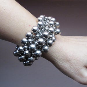 May include: A silver bracelet made of many small, spherical beads. The bracelet is worn on a wrist, with the beads clustered together to create a textured, striking accessory. The background is a soft, neutral tone.