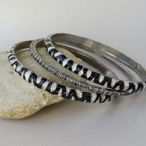 May include: Three silver bangles. Two feature a black and white zigzag design, and the third is decorated with small, clear crystals. The bangles are stacked on a light-coloured stone.