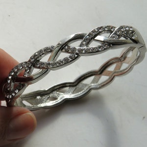 May include: Silver-toned bracelet with a braided design, embellished with clear rhinestones. The bracelet is approximately 2.5 inches in diameter and features an intricate, interwoven pattern. A hinged clasp secures the bracelet.