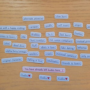 May include: A collection of white rectangular stickers with various phrases related to romantic relationships, such as "enemies to lovers", "slow burn", "soulmates", and "friends to lovers". The stickers are arranged on a wooden surface with three red heart stickers at the bottom.