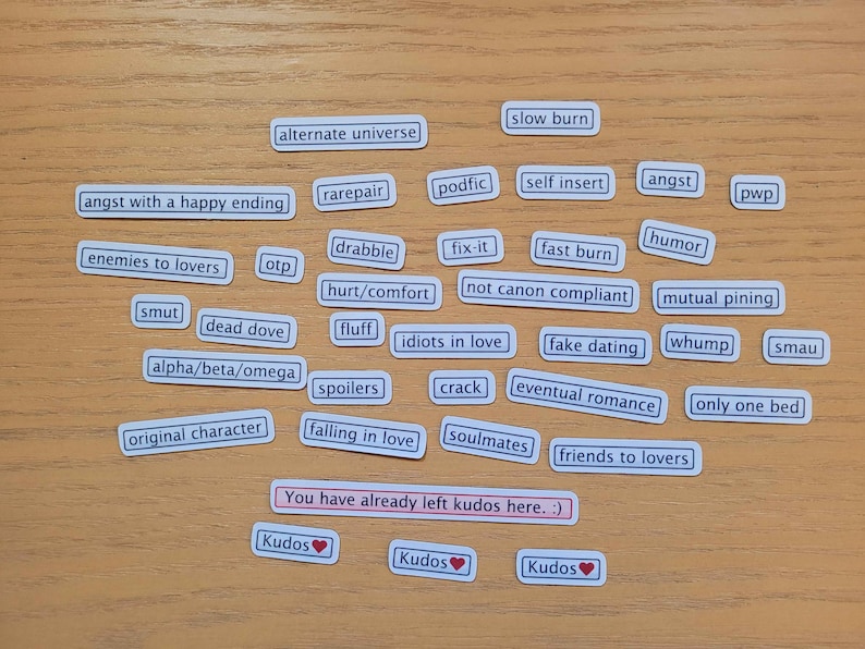 May include: A collection of white rectangular stickers with various phrases related to romantic relationships, such as "enemies to lovers", "slow burn", "soulmates", and "friends to lovers". The stickers are arranged on a wooden surface with three red heart stickers at the bottom.