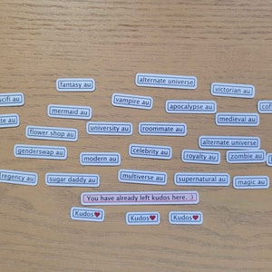 May include: A collection of white rectangular stickers with black text on a wooden surface. The stickers feature various alternate universe (AU) prompts for creative writing, including "fantasy au", "scifi au", "vampire au", "apocalypse au", "soulmate au", "mermaid au", "flower shop au", "genderswap au", "regency au", "sugar daddy au", "university au", "roommate au", "modern au", "celebrity au", "multiverse au", "victorian au", "medieval au", "alternate universe", "royalty au", "zombie au", "supernatural au", "coffee shop au", "alien au", "band au", and "magic au".