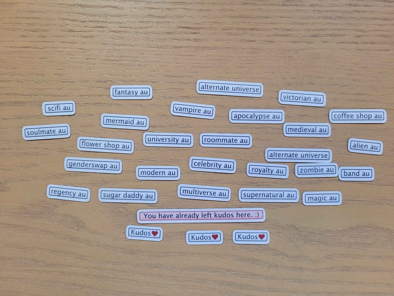 May include: A collection of white rectangular stickers with black text on a wooden surface. The stickers feature various alternate universe (AU) prompts for creative writing, including "fantasy au", "scifi au", "vampire au", "apocalypse au", "soulmate au", "mermaid au", "flower shop au", "genderswap au", "regency au", "sugar daddy au", "university au", "roommate au", "modern au", "celebrity au", "multiverse au", "victorian au", "medieval au", "alternate universe", "royalty au", "zombie au", "supernatural au", "coffee shop au", "alien au", "band au", and "magic au".