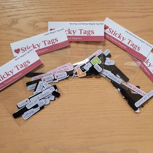May include: Four clear plastic bags containing sets of black magnetic tags with various phrases and words printed on them. The bags are labelled "Sticky Tags" and each set has a different theme: "Basics", "Fanfiction", "Warnings and Ratings", and "Alternate Universes".