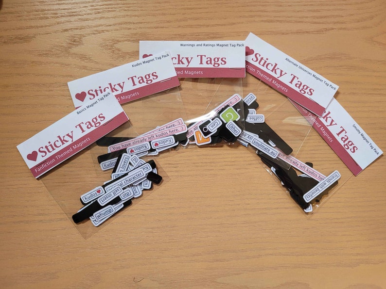 May include: Four clear plastic bags containing sets of black magnetic tags with various phrases and words printed on them. The bags are labelled "Sticky Tags" and each set has a different theme: "Basics", "Fanfiction", "Warnings and Ratings", and "Alternate Universes".