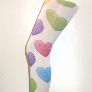 May include: White tights with colorful candy heart designs. The hearts have messages like "Kiss Me," "Call Me," and "Miss You." The tights are displayed on a metal stand.