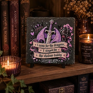 Peut inclure: Une plaque en ardoise carrée ornée d'une épée violette avec des ailes de dragon, une couronne et un casque de chevalier. Une bannière en ruban rose affiche le texte : I came for the dragons, I stayed for the shadow daddy, et romantasy girlie.