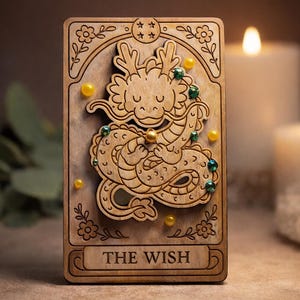 Peut inclure: Carte de tarot en bois avec un motif de dragon orné de décorations jaunes et vertes. La carte porte l'inscription "THE WISH" en bas. Le dragon est finement sculpté et entouré d'éléments floraux. L'arrière-plan comprend des bougies allumées et du feuillage.
