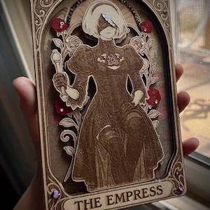 Peut inclure: Carte de tarot en bois représentant une figure en robe longue tenant un bâton, avec le texte "THE EMPRESS". La carte présente un motif floral avec des roses rouges et des accents décoratifs.