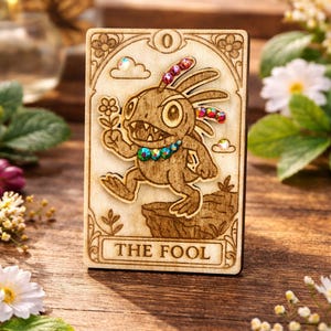 Peut inclure: Carte de tarot en bois représentant une créature de dessin animé tenant une fleur, ornée de pierres précieuses colorées. La carte porte l'inscription "THE FOOL". Le fond est en bois.