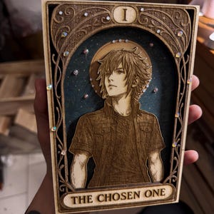 Puede incluir: Obra de arte de madera que presenta un retrato de un personaje con pelo puntiagudo, sobre un fondo estrellado. La obra está enmarcada con elementos decorativos y el texto "THE CHOSEN ONE". Adornada con cristales iridiscentes.
