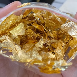 May include: A transparent plastic bag containing gold leaf flakes. The flakes are metallic, with an uneven, textured appearance. The bag is held in a hand, with a blurred background.