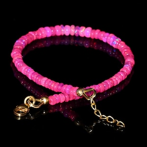 Peut inclure: Un bracelet de perles roses avec un fermoir et une chaîne dorés. Le bracelet est composé de petites perles roses rondes. Le fermoir et la chaîne sont dorés. Le bracelet est présenté sur une surface noire.