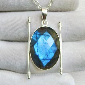 May include: A silver pendant necklace featuring a large, oval-shaped, faceted labradorite gemstone with a vibrant blue flash. The stone is set in a silver frame with two vertical silver bars on either side, suspended from a delicate silver chain.