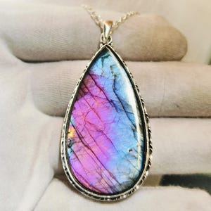 May include: A teardrop-shaped pendant necklace featuring a polished labradorite gemstone with vibrant iridescent flashes of purple, blue, and gold. The stone is set in a silver-toned metal frame with a textured edge, suspended from a silver chain.