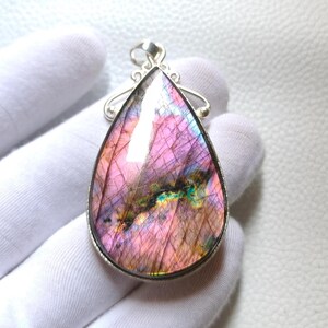 May include: A teardrop-shaped labradorite pendant set in a silver-toned metal frame. The gemstone displays iridescent pink, purple, and blue hues with internal fractures and a prominent dark green and gold inclusion across the center.