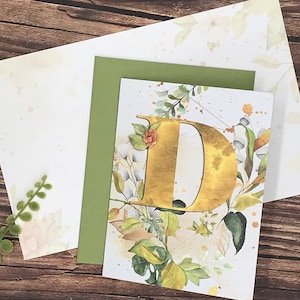May include: A white card with a gold letter D surrounded by watercolor flowers and leaves. The card is on a green background.