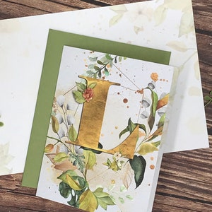 May include: A white card with a gold letter L surrounded by green and white floral illustrations. The letter L is surrounded by a geometric pattern with gold accents.