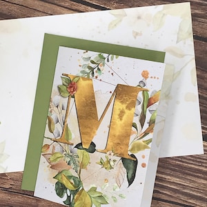 May include: A white card with a gold letter "M" surrounded by watercolor floral illustrations. The card is accented with a green border.