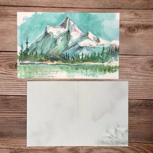 K&ouml;nnte beinhalten: Ein Aquarellbild einer Bergkette mit einem See im Vordergrund. Die Berge sind schneebedeckt und der See ist hellblau. Das Bild ist auf einer wei&szlig;en Karte mit dem Text "Little Wren Card Company" und einer Website-Adresse.