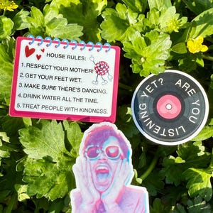 May include: A collection of stickers on a bed of green foliage. One sticker is a notepad with "HOUSE RULES" and bullet points. Another is a circular sticker with a record design and the words "ARE YOU LISTENING YET?". The third sticker is a colorful image of a person.